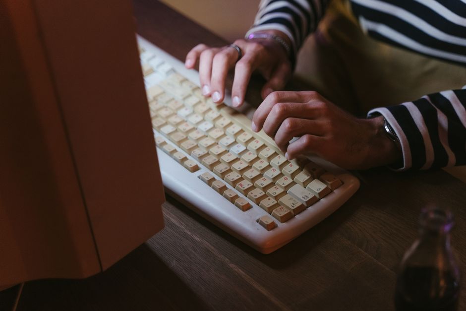 Dos manos tecleando en un teclado beige de los de antes, en penumbra, con una botella de refresco al lado.
